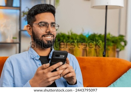 Happy young man thinking and text messaging on smartphone. Indian guy sitting on sofa and using cellphone in living room at home. Arabian male is searching information on browser internet. Copy-space