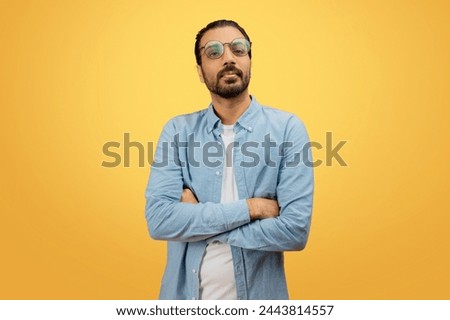 A self-assured indian man with a beard and glasses stands with crossed arms in a denim shirt, against a yellow backdrop Royalty-Free Stock Photo #2443814557