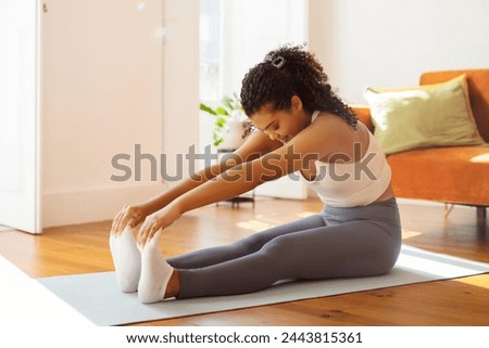 Millennial fit woman in living room sitting on mat in sportswear bending touching toes, follows fitness routine improving flexibility, emphasizing wellbeing and healthy lifestyle habits Royalty-Free Stock Photo #2443815361