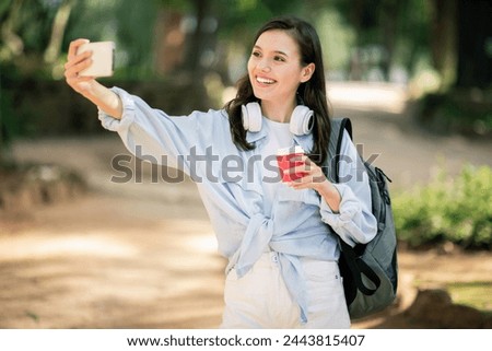 Vivacious happy young european woman taking a selfie with her smartphone, holding a red coffee cup, with headphones around her neck, enjoy device, in a sunny park setting, outside Royalty-Free Stock Photo #2443815407