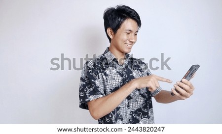 Handsome young Asian man wearing batik is making a video call, taking a selfie using a smartphone on an isolated white background