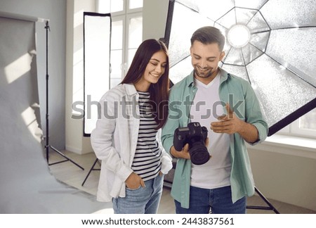 Portrait of a young smiling professional photographer showing photos to a girl model on digital camera standing in production studio with light equipment and discussing photo session.