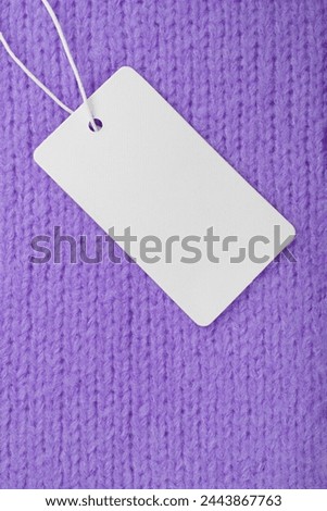 Blank clothing label on purple knitted sweater background. Mockup, template with empty rectangular card, cloth tag for price or text. Shopping, sale, discount, black friday, fashion concept.