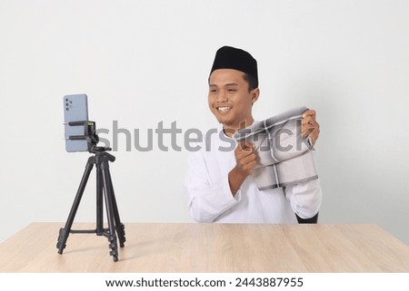 Portrait of excited Asian muslim man in koko shirt with skullcap promoting his product, sarung, on live streaming session. Online shopping with smartphone concept. Isolated image on white background