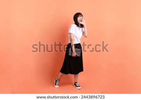 Cheerful female student in Thai university uniform with gesture of holding a textbook isolated on orange background. International Students' Day, World Students' Day. Gen Z
