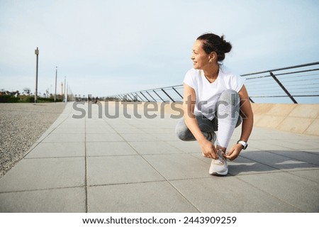 Young woman jogger tying laces on her pink sneakers, smiling looking away, getting ready for morning jog in the city outdoors. Running and jogging. Awareness running marathon concept. Sport. Endurance Royalty-Free Stock Photo #2443909259