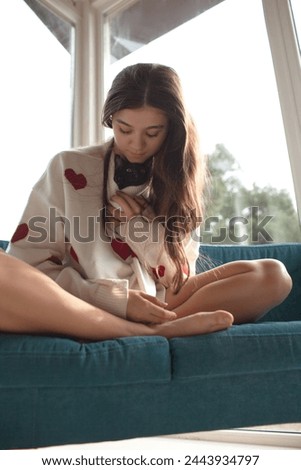 The girl holding the black cat. Friendship between the human and animal. The female dressed the white sweater with red heart. The portrait of young woman holding the kitten lies on sofa