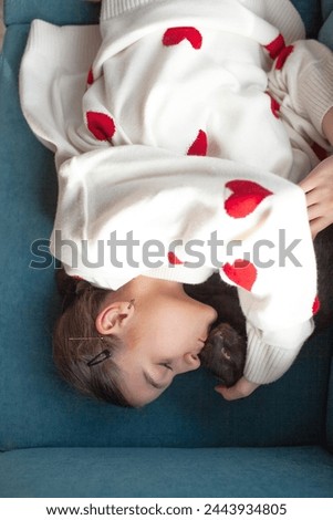 The girl holding the black cat. Friendship between the human and animal. The female dressed the white sweater with red heart. The portrait of young woman holding the kitten lies on sofa