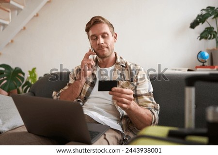 Portrait of young man with credit card, sitting and using laptop, making a phone call, booking hotel or tickets, confirming his purchase over telephone, has suitcase near him. Royalty-Free Stock Photo #2443938001