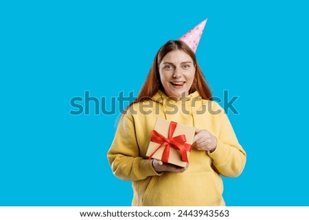 Young happy 30s woman in birthday party hat holding present on blue background. Emotional, young face. Human emotions, facial expression and holiday concept.