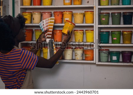 Smiling multicultural graphic technologist standing at printing workshop with color swatch and bucket of paint in her hands and matching colors. Rear view of african american worker choosing colors.
