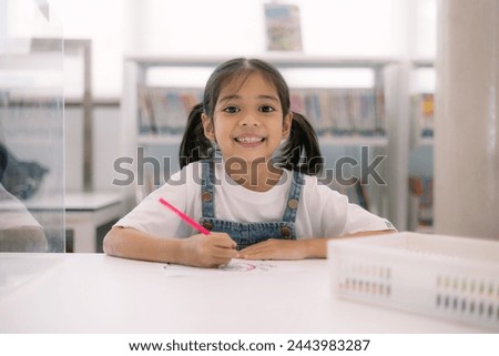 A young girl is sitting at a desk with a red crayon in her hand. She is smiling and she is enjoying herself. The scene suggests a positive and playful atmosphere Royalty-Free Stock Photo #2443983287