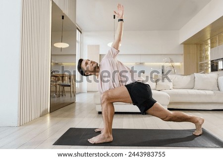 A man performs a yoga stretch with an extended arm, focusing on his form and balance in a spacious, contemporary living area.

 Royalty-Free Stock Photo #2443983755