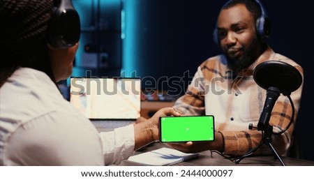 Man invited to podcast watching internet clips on green screen smartphone with host in studio, discussing content. BIPOC guest reacting to videos on mockup mobile phone during online comedy show