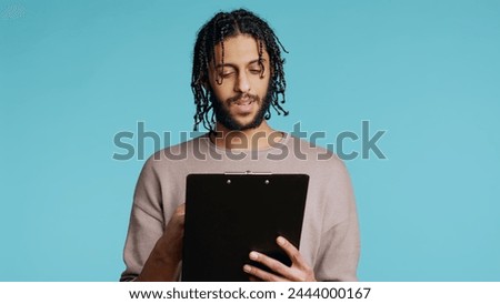 Public speaker reading from clipboard files, preparing discourse, rehearsing before doing speech. Middle Eastern orator telling monologue, isolated over studio background, camera B Royalty-Free Stock Photo #2444000167