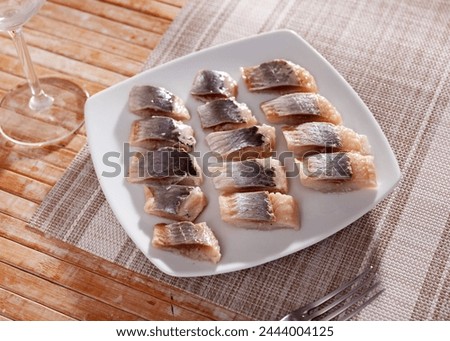 On table is plate with appetizer - pieces of salted herring fillet. Gentle refection for light alcoholic beverages. Fish supplemented with edible green Royalty-Free Stock Photo #2444004125