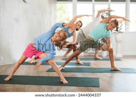 Family with children performs physical education at home lateral elongated angle pose, Parshvakonasana. Little confident girl with older brother and parents does balance asana from yoga practice