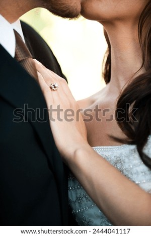Closeup of the wedding ring of a newly married bride. Her hand is on her grooms chest and is framed by the bride and groom kissing. It is a unique wedding ring.  Royalty-Free Stock Photo #2444041117