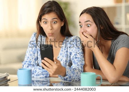 Two amazed women checking smart phone at home