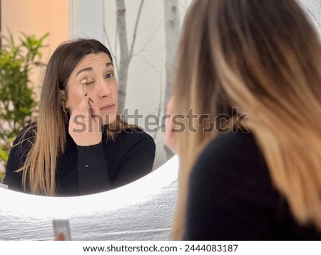 Woman in black attire applying eye makeup, reflected in a vanity mirror.