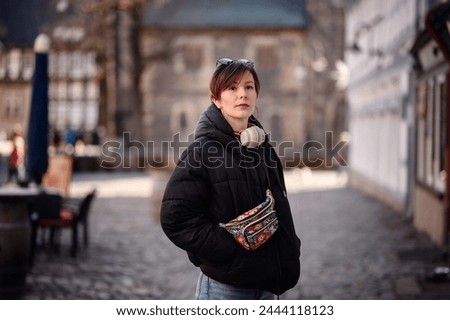 A poised adult woman with short hair and red lipstick stands confidently outdoors. She sports casual attire with a patterned fanny pack and headphones around her neck,  Royalty-Free Stock Photo #2444118123