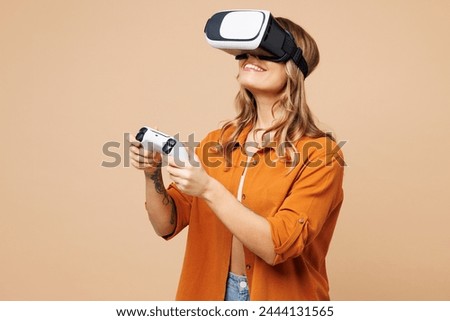 Young woman wear orange shirt casual clothes watching in vr headset pc gadget hold play pc game with joystick console isolated on plain pastel light beige background studio portrait. Lifestyle concept