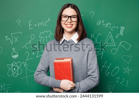 Young fun smiling smart teacher woman wear grey casual shirt glasses hold in hand book look camera isolated on green wall chalk blackboard background studio. Education in high school college concept