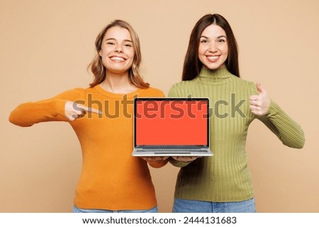 Young friends two IT women wear orange green shirt casual clothes together hold use work on laptop pc computer show thumb up isolated on plain pastel light beige background studio. Lifestyle concept Royalty-Free Stock Photo #2444131683
