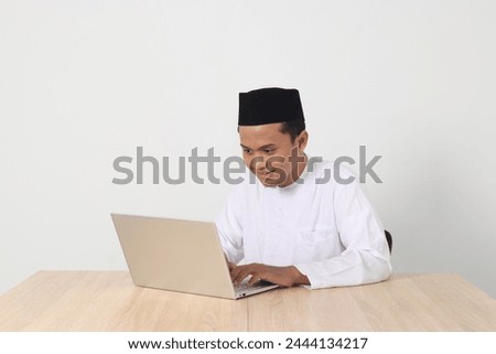 Portrait of excited Asian muslim man in koko shirt with skullcap working on his laptop during fasting on ramadan month. Isolated image on white background