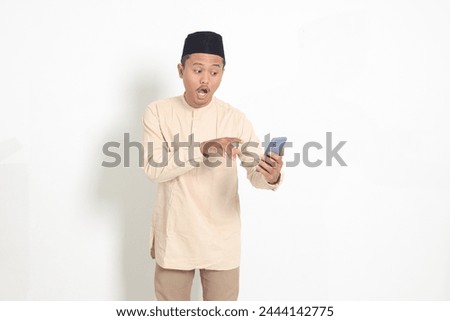 Portrait of surprised Asian muslim man in koko shirt with skullcap holding mobile phone, showing shocked face expression. Advertising and social media concept. Isolated image on white background
