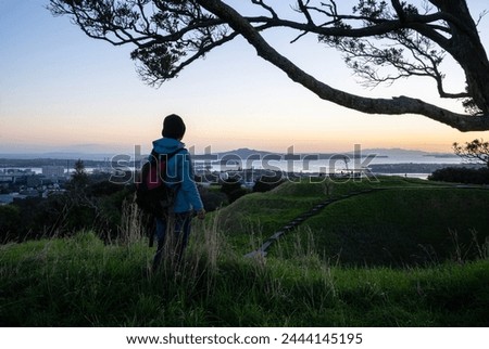 Woman enjoying the sunrise at Mt Eden summit with volcanic crater in the foreground. Rangitoto Island framed by Pohutukawa trees. Auckland. Royalty-Free Stock Photo #2444145195