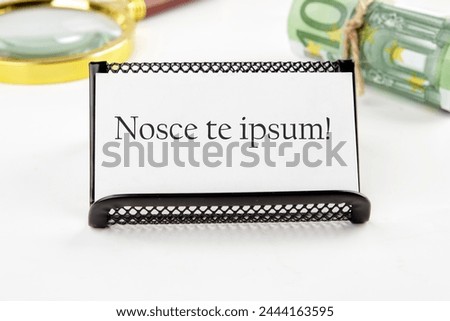 Latin proverb NOSCE TE IPSUM (know yourself) on a white business card on a white background in a black stand next to it in non-focus are money and a magnifying glass