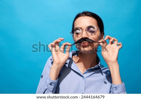 Young woman in shirt, wearing fake moustaches, glasses with eyes items on it and making funny expressions against blue background. Concept of food pop art photography, creativity, quirky style