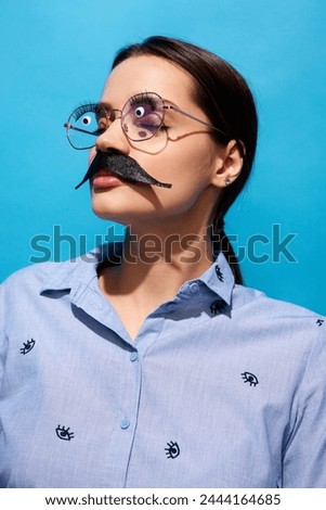 Young woman in shirt, wearing fake moustaches, glasses with eyes items on it standing with closed eyes against blue background. Concept of food pop art photography, creativity, quirky style