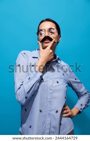 Young woman in shirt, wearing fake moustaches, glasses with eyes items on it and making funny expressions against blue background. Concept of food pop art photography, creativity, quirky style