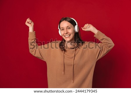 Cheerful young woman wearing headphones is dancing over red background.