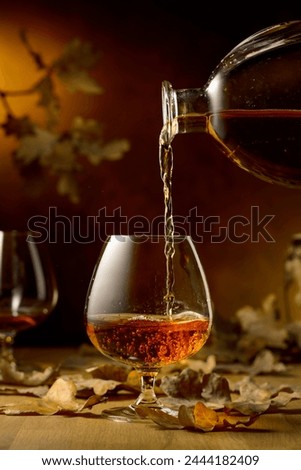 Cognac is poured into a glass from a bottle. A snifter of brandy on an old oak table with fallen oak leaves. Royalty-Free Stock Photo #2444182409