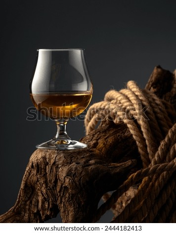 Brandy snifter and rope on a old wooden snag. Glass with whiskey, cognac or brandy on a dark background. Royalty-Free Stock Photo #2444182413