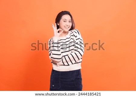 Portrait of a beautiful cheerful confident indonesia girl standing with arms crossed gesturing okay good good and looking at the camera isolated on an orange background wearing a white sweater