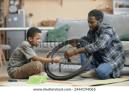 Side view portrait of African American father and son repairing bicycle wheel together sitting on floor at home