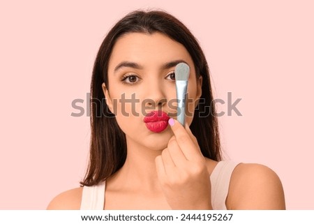 Beautiful young woman with stylish makeup and brush blowing kiss on pink background