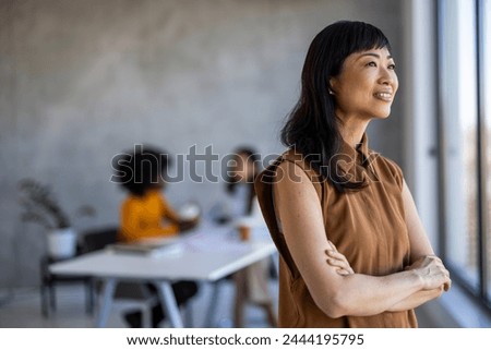 Portrait of successful young Asian businesswoman at office, colleagues in background. Smiling confident Asian businesswoman with arms crossed standing in modern office with colleagues.