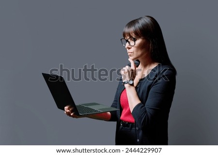 Serious middle aged woman holding laptop on gray background