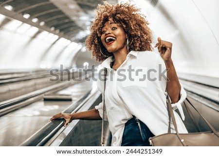 Beautiful black woman standing on escalator on her way to modern brightly lit subway station. Public transportation and urban life concept. Low angle shot. Royalty-Free Stock Photo #2444251349