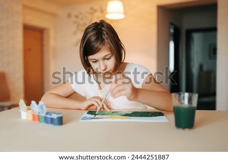 Child girl sits at the table and draws a picture in the album while looking at her. Both hands on the table, focused look light clothes. Near a glass of water and paint. Front view.