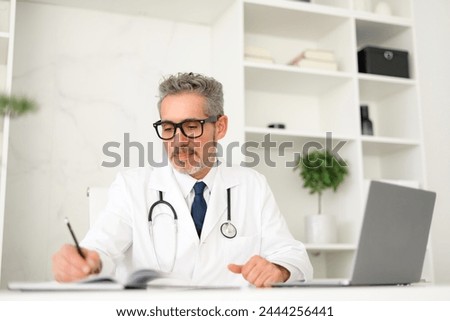 Focused grey-haired doctor taking notes while using laptop, reviewing patient files or medical literature. His concentration reflects diligence and expertise expected in modern medical care Royalty-Free Stock Photo #2444256441