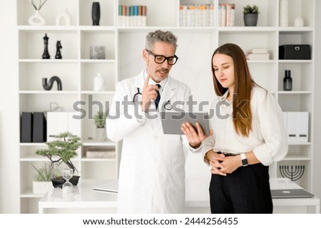 A grey-haired male doctor in a white coat discusses medical information with a young woman, using a tablet to enhance understanding, standing in a modern office Royalty-Free Stock Photo #2444256455
