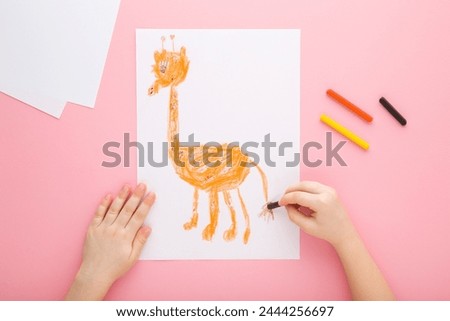 Little child girl hand holding wax crayon and drawing orange cute giraffe shape on white paper on light pink table background. Pastel color. Closeup. Point of view shot. Children creativity time.