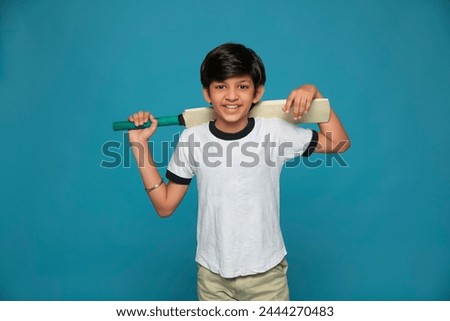 Portrait of smiling boy standing with cricket bat on shoulder against blue background