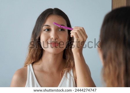 Dermaplaning. Pretty hispanic girl shaving her eyebrows by razor on the mirror at home. Facial hair removal.